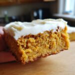 A close-up shot of a moist pumpkin bar topped with creamy cream cheese frosting.