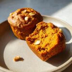 Close-up of two pumpkin protein muffins on a plate, one whole and one broken in half, showing a moist interior with nuts.