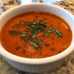 A close-up of a bowl of creamy roasted tomato basil soup, garnished with fresh basil leaves.
