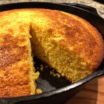 Close-up of golden cornbread baked in a cast-iron skillet, with a slice removed.