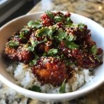 A bowl of fluffy coconut rice topped with glistening spicy maple chicken pieces, garnished with sesame seeds and cilantro.