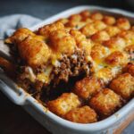 A serving spoon lifts a portion of Tater Tot Casserole, revealing layers of ground beef and melted cheese topped with golden tater tots.