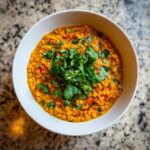 A close-up overhead shot of a white bowl filled with vibrant orange lentil curry, garnished with fresh cilantro.