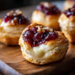 Close-up of a warm brie bite showing melted brie cheese inside flaky pastry topped with cranberry sauce and walnuts.