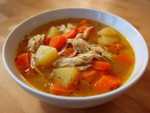 A close-up of a white bowl filled with rich, golden chicken vegetable soup featuring shredded chicken, carrots, and potatoes.