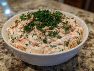 A white bowl filled with creamy, pink salmon dip generously topped with fresh dill.