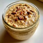 Close-up of apple cinnamon overnight oats in a glass jar, topped with chopped pecans and cinnamon.