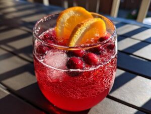 A close-up of a glass of autumn punch, featuring a vibrant red liquid, ice cubes, whole cranberries, and orange slices.
