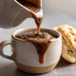 Close-up of black pepper cream gravy being poured from a creamer into a rustic mug, with a biscuit in the background.