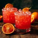 Two glasses of vibrant red blood orange margarita cocktails, garnished with orange slices and rimmed with salt, on a wooden table.