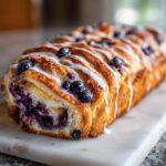 Close-up of a golden-brown Blueberry Lemon Crescent Ring, drizzled with white icing and topped with fresh blueberries.