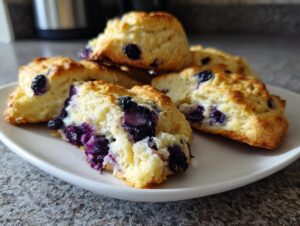 Close-up of flaky blueberry scones piled on a white plate, showcasing juicy blueberries within.