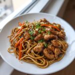 A close-up of a white plate filled with savory chicken chow mein recipe, featuring saucy chicken pieces, noodles, carrots, and green onions.