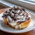 A close-up of a warm, gooey cookies and cream cinnamon roll topped with white frosting and crushed cookies.