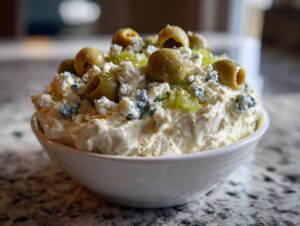 A close-up of a creamy dirty martini dip topped generously with blue cheese crumbles, pimento-stuffed green olives, and olive brine pieces.