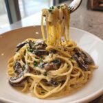 A fork lifting strands of creamy mushroom pasta from a white bowl, showing rich sauce and sliced mushrooms.