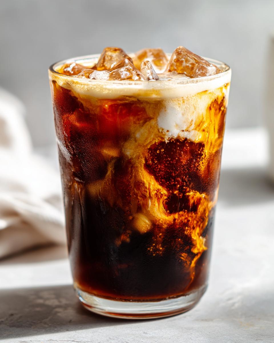 A tall glass of New Orleans style crème brûlée iced coffee with ice cubes and swirling cream.