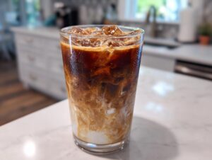 A tall glass filled with New Orleans style crème brûlée iced coffee, showing swirls of cream and dark coffee over ice.