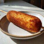 A single, perfectly golden brown and crispy corn dog served on a small, light-colored plate.