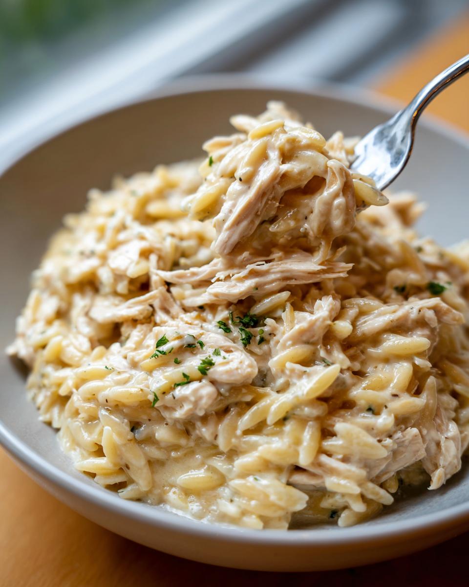 A fork lifting a bite of creamy Crockpot Chicken Alfredo Orzo from a bowl, garnished with parsley.