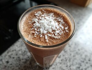 A refreshing dark chocolate coffee smoothie topped with shredded coconut flakes in a clear glass.