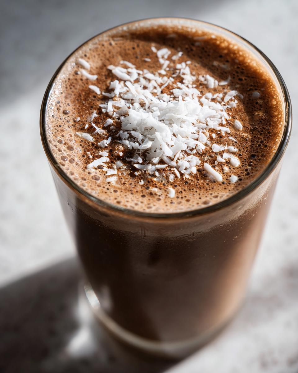 A close-up of a dark chocolate coffee smoothie topped with shredded coconut flakes in a clear glass.