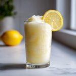 A tall glass filled with icy Frozen Creamy Lemonade, garnished with a lemon slice.
