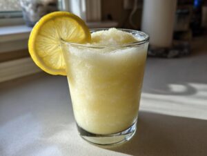 A close-up of a tall glass filled with icy Frozen Creamy Lemonade, garnished with a lemon slice.