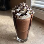A tall glass of rich, blended frozen hot chocolate topped with whipped cream, chocolate syrup, and shavings.