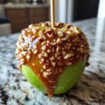 A close-up photo of a bright green caramel apple dipped in gooey caramel and covered in chopped pecans.