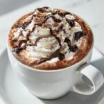 A close-up of a steaming mug of hazelnut hot chocolate topped with whipped cream, chocolate drizzle, and shaved hazelnuts.