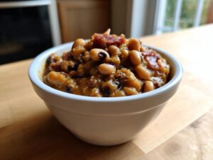 A close-up of a white bowl filled with savory black eyed peas stewed in a rich sauce and topped with bacon pieces.