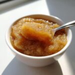 Close-up of chunky, homemade applesauce in a white bowl, with a spoonful being lifted out, showing cinnamon specks.