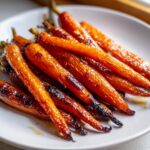 A close-up of perfectly roasted, glossy honey glazed carrots piled on a white plate.