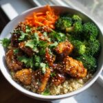 A close-up of a hot honey chicken bowl featuring glazed chicken pieces, steamed broccoli, shredded carrots, and quinoa, garnished with sesame seeds and cilantro.