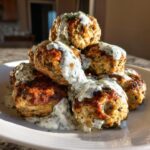 A stack of golden brown greek chicken meatballs generously drizzled with creamy white tzatziki sauce and fresh dill.