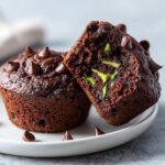 Close-up of two moist chocolate zucchini muffins, one cut in half showing green zucchini shreds inside.