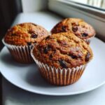 Close-up of three freshly baked, moist bran muffins studded with raisins, resting on a white plate.