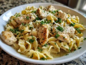 A close-up of a white bowl filled with one-pan chicken and buttered noodles, garnished with parsley and Parmesan cheese.