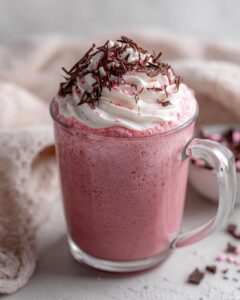 A festive glass mug filled with frothy ruby hot chocolate, topped with whipped cream and dark chocolate shavings.