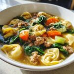 A close-up view of a steaming bowl of sausage tortellini soup, featuring tortellini, ground sausage, spinach, and carrots.