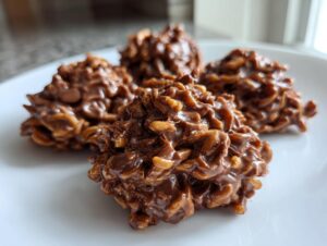Four glossy, chocolate-covered haystack cookies made with cereal and nuts, sitting on a white plate.