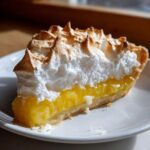 A close-up of a slice of lemon meringue pie showing bright yellow filling and tall, toasted meringue peaks.