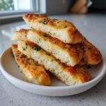 A stack of soft, golden-brown breadsticks topped with garlic, cheese, and parsley served on a small white plate.