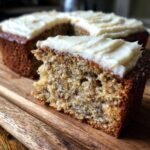 A slice of moist sourdough banana cake with creamy white frosting on a wooden board.