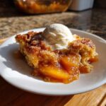 A warm slice of southern peach cobbler topped with melting vanilla ice cream on a white plate.