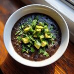 A close-up of a bowl of thick, dark black bean soup garnished with diced avocado and fresh cilantro.