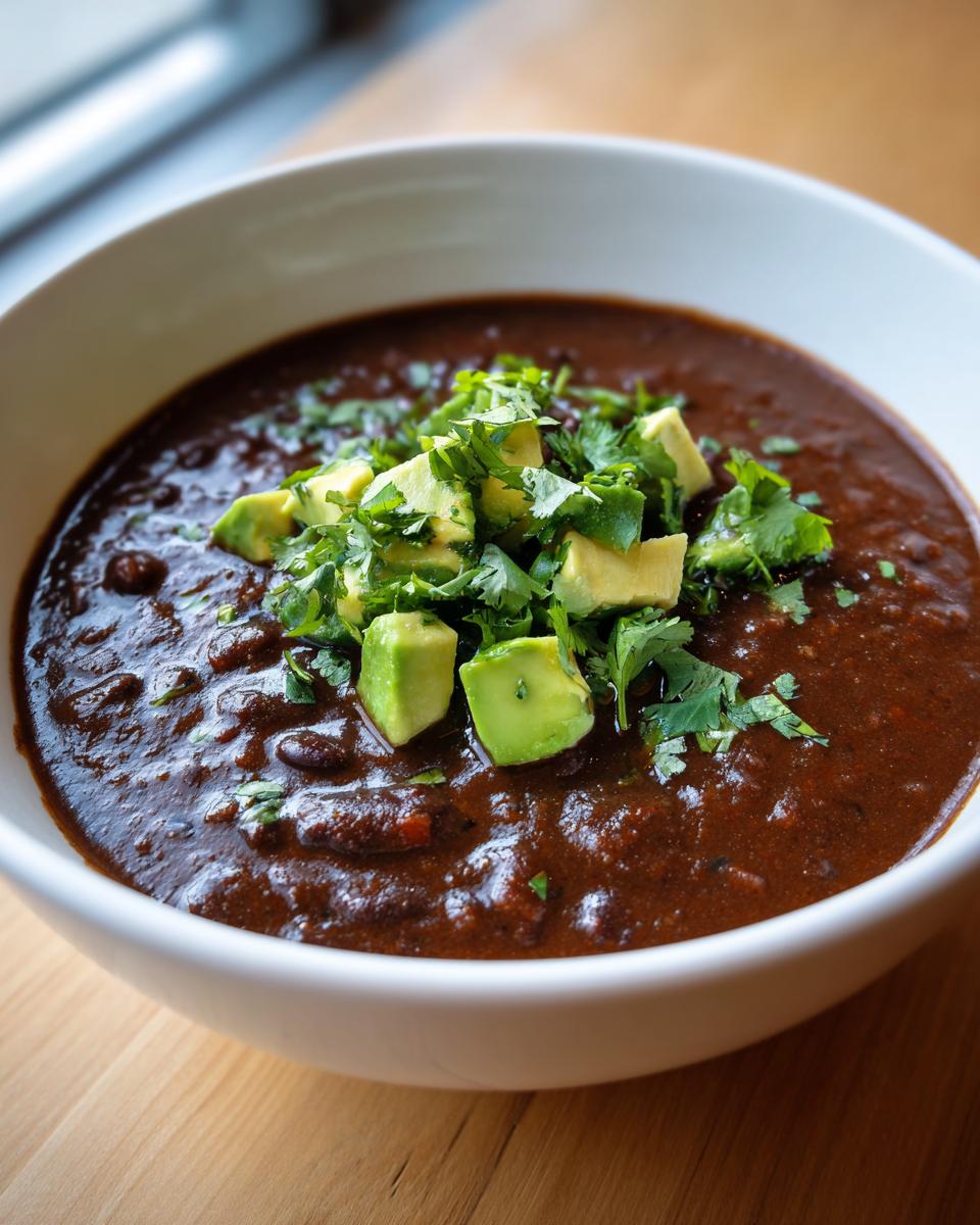 Close-up of a bowl of rich, dark spicy black bean soup garnished with diced avocado and fresh cilantro.