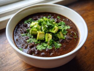 A close-up of a hearty bowl of spicy black bean soup, garnished generously with diced avocado and fresh cilantro.