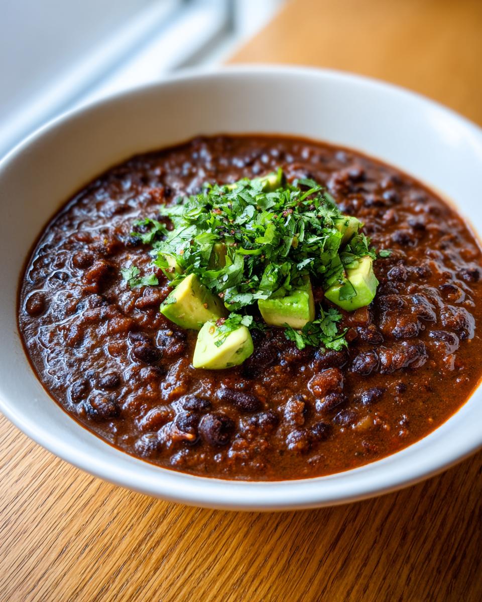 Close-up of a bowl of rich, dark spicy black bean soup garnished with diced avocado and fresh cilantro.